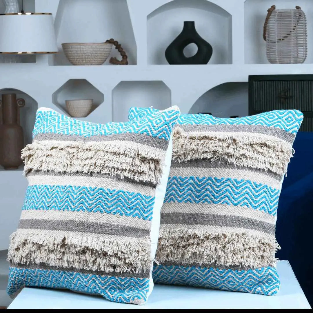 Set of two decorative boho pillow covers with blue chevron design, beige tufted fringe, and gray woven stripes, displayed on a modern blue chair in a stylish living room.