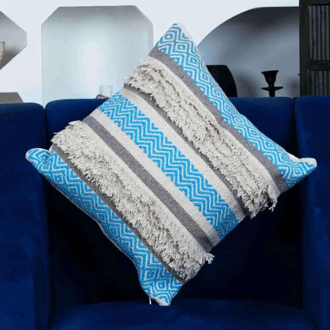 Boho throw pillow cover with blue chevron pattern, beige tufted fringe, and gray woven stripes, displayed on a navy blue armchair in a modern living room.