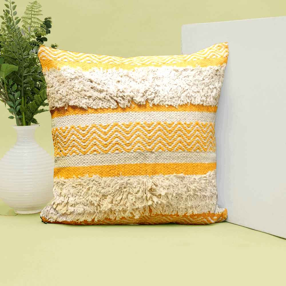 Yellow woven tufted throw pillow with beige fringe and zigzag pattern, displayed upright against a white panel with a ceramic white vase and greenery beside it on a pastel green surface.