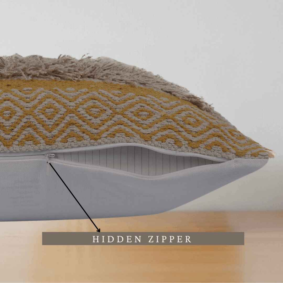 Close-up side view of a decorative mustard yellow and beige woven tufted pillow, highlighting a hidden zipper opening at the bottom, with textured fabric and wooden surface underneath.