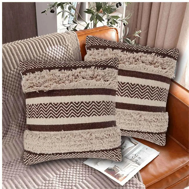 Two brown and beige decorative throw pillows with zigzag woven tufted patterns and fringe detailing, styled on a tan leather sofa with a textured throw blanket and a magazine.