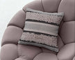 Grey and black woven textured throw pillow with tufted fringes and zigzag patterns, styled on a modern mauve upholstered chair for a cozy and contemporary home aesthetic.