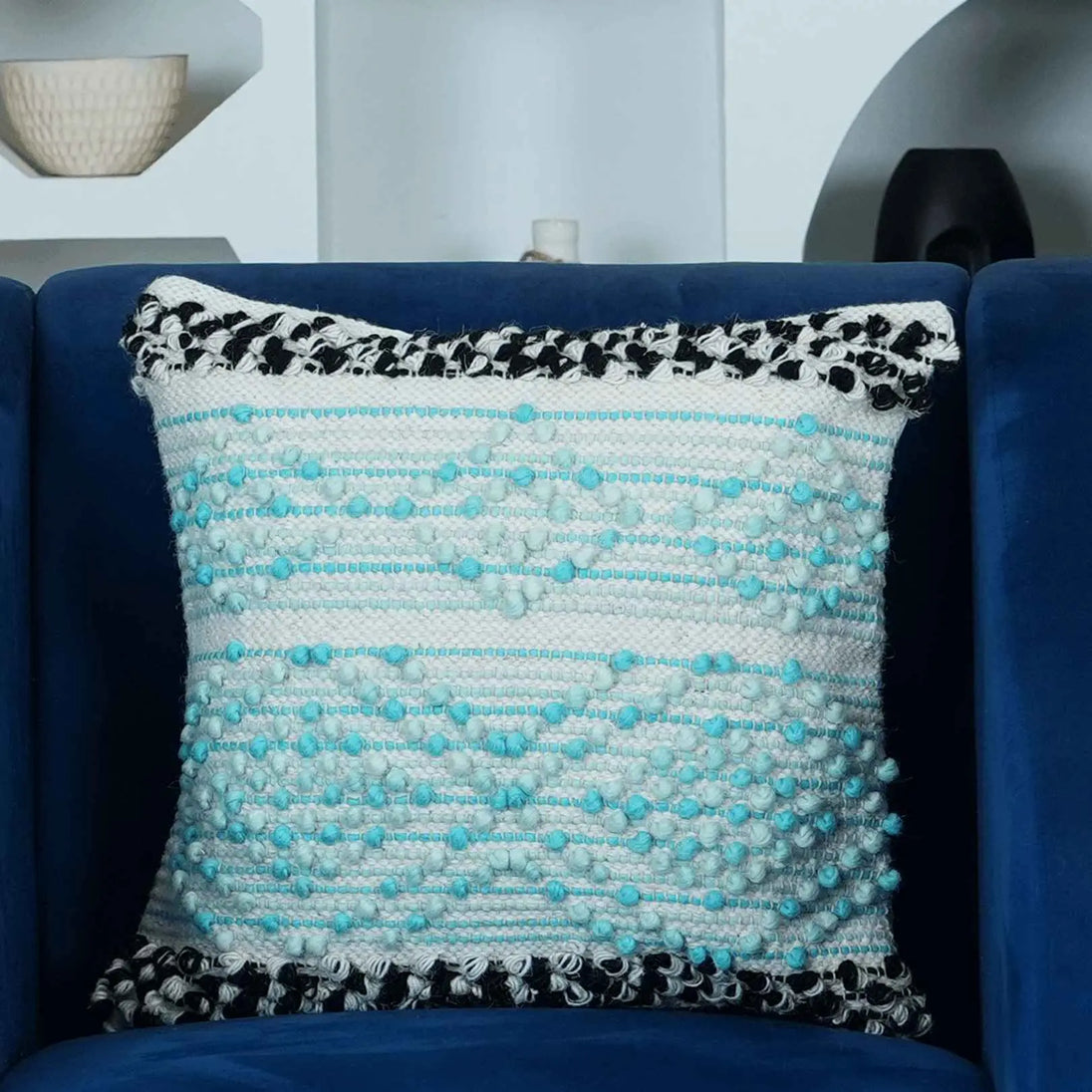Textured boho throw pillow cover in aqua blue and white with black tufted edges, displayed on a navy blue armchair in a stylish modern living room.







