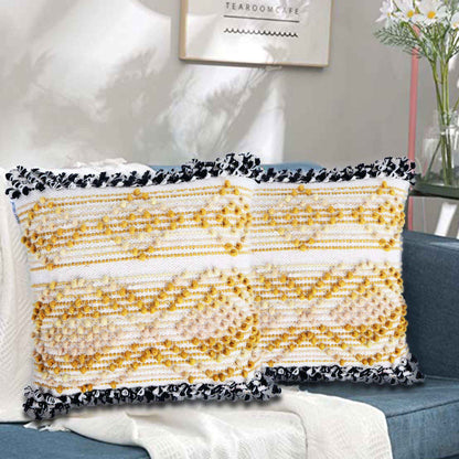 Two decorative yellow and white tufted cushion covers with black-and-white tassel edges placed on a blue sofa. The cushions feature a textured, geometric woven pattern, with a cozy cream throw draped beneath them. The setting is a modern living room with wall art, indoor plants, and soft natural lighting.









