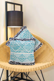 Set of boho throw pillow covers in aqua blue and white with black fringe accents, displayed on a woven rattan chair beside a modern floor lamp in a cozy home interior.







