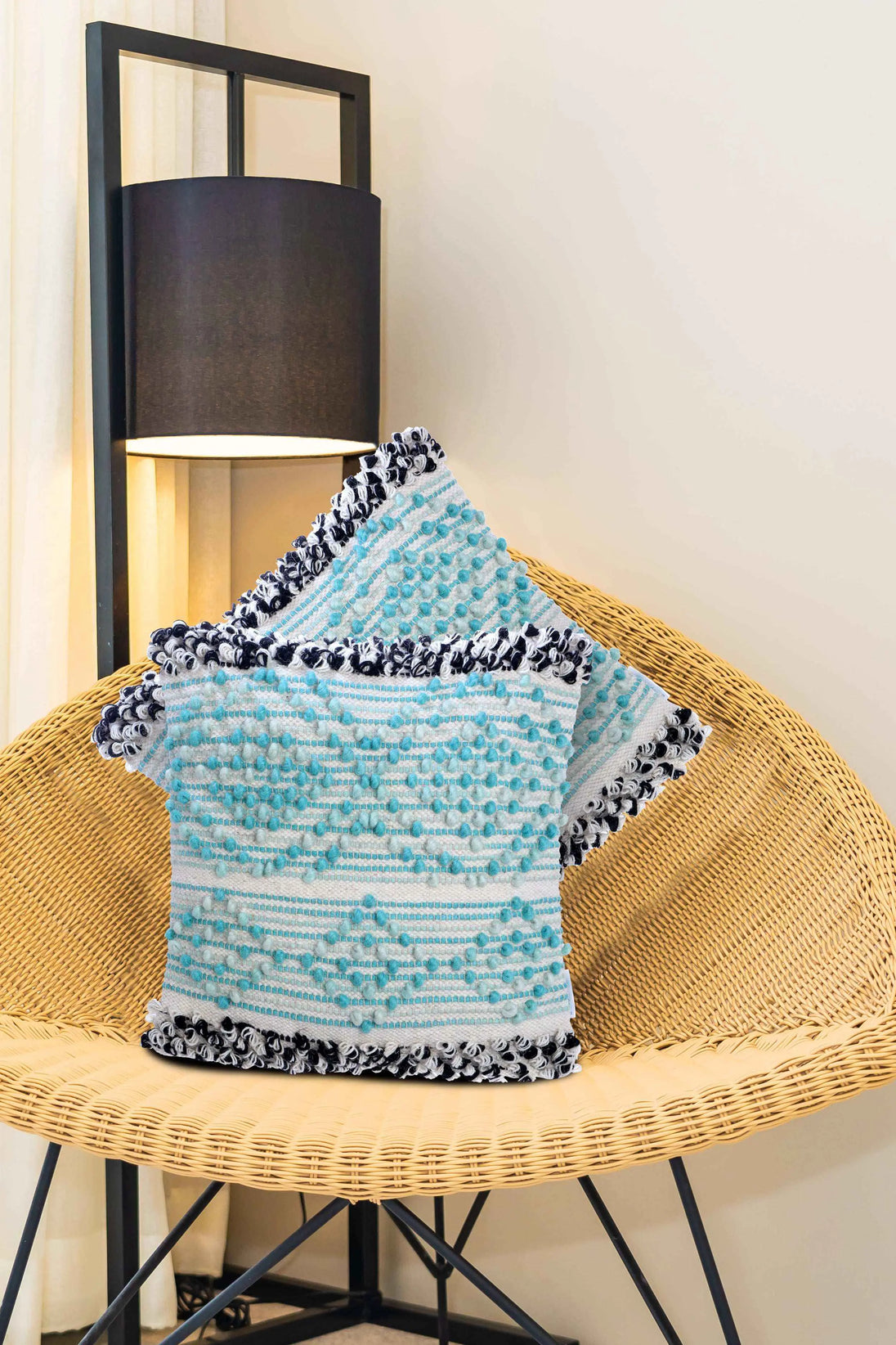 Set of boho throw pillow covers in aqua blue and white with black fringe accents, displayed on a woven rattan chair beside a modern floor lamp in a cozy home interior.







