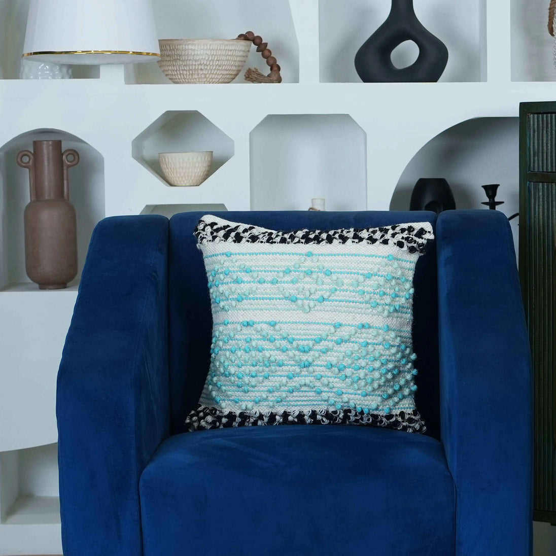 Boho textured pillow cover in aqua blue and white with black tufted edges, styled on a modern navy blue accent chair in a contemporary living room setting.







