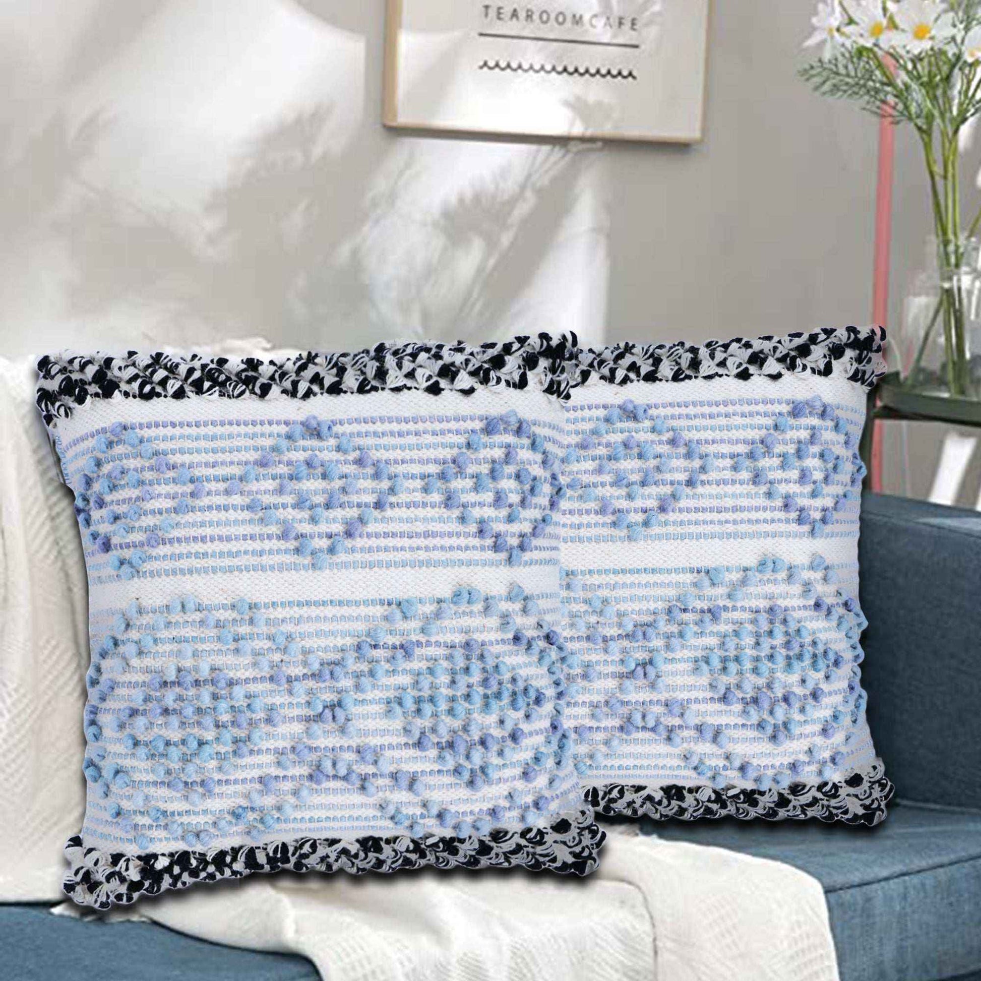 Two decorative square cushion covers with a soft blue and white tufted pattern and black-and-white braided edges, placed on a cozy couch in a modern, sunlit living room with a minimalist decor vibe.







