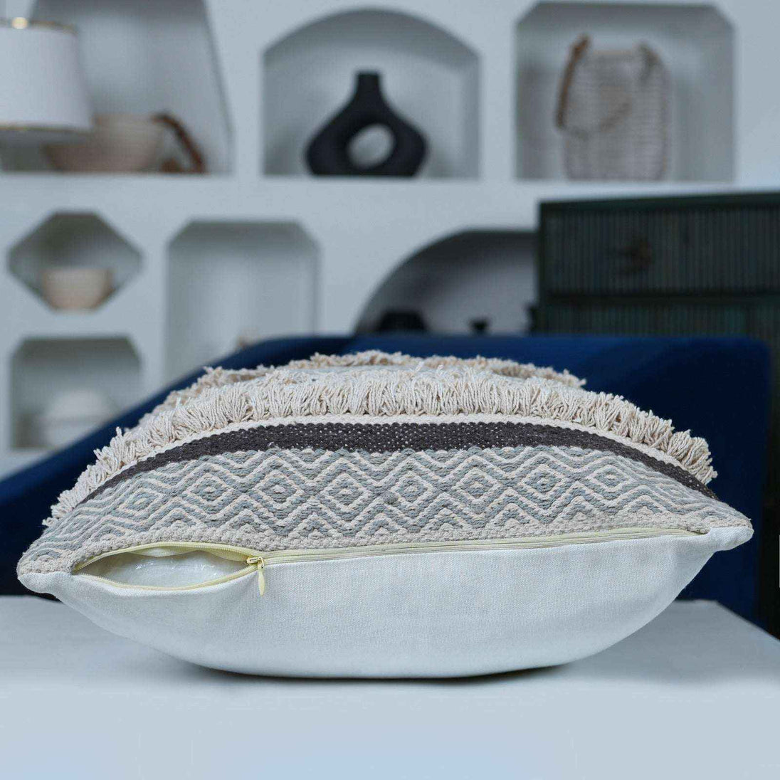 Side profile of a textured throw pillow with a beige and grey woven tufted front, featuring a partially open hidden zipper at the bottom revealing the inner pillow insert, placed on a modern white surface with decorative shelving in the background.







