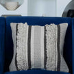 Decorative woven throw pillow cover with textured zigzag and tufted fringe patterns in grey and black tones, placed on a royal blue armchair in a modern living room setting.