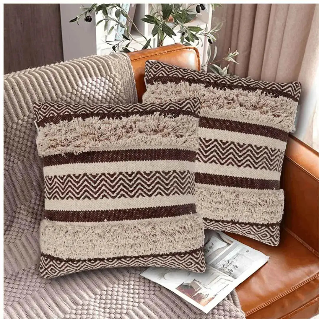 Two brown and beige decorative throw pillows with zigzag woven tufted patterns and fringe detailing, styled on a tan leather sofa with a textured throw blanket and a magazine.