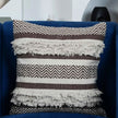 Brown and cream woven tufted throw pillow with fringe detailing and zigzag pattern, displayed on a dark blue velvet chair in a modern living room setup.








