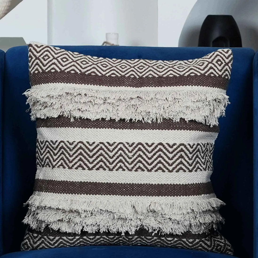 Brown and cream woven tufted throw pillow with fringe detailing and zigzag pattern, displayed on a dark blue velvet chair in a modern living room setup.







