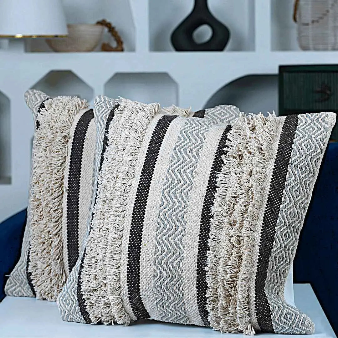 Two decorative square pillows with tufted woven texture in cream, grey, and black chevron and stripe patterns, placed on a modern white surface in front of a blue armchair and a white wall with geometric shelves in the background.