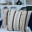 Two decorative square pillows with tufted woven texture in cream, grey, and black chevron and stripe patterns, placed on a modern white surface in front of a blue armchair and a white wall with geometric shelves in the background.