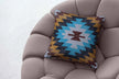 Southwestern tribal print throw pillow with bold geometric patterns in turquoise, mustard yellow, maroon, and black, placed on a modern tufted beige armchair, creating a cozy and stylish accent.