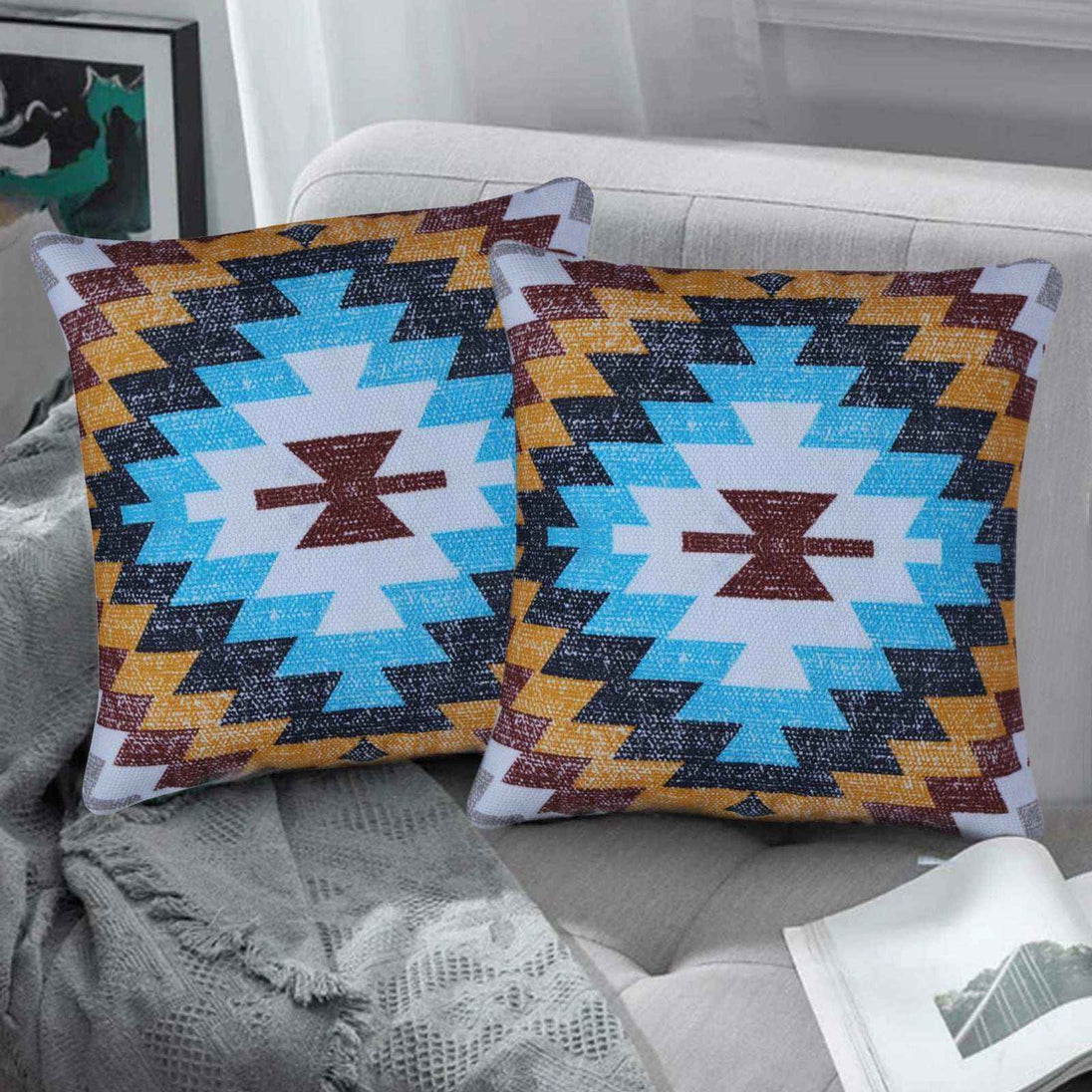 Set of two tribal geometric throw pillows featuring a bold Southwestern pattern in turquoise, mustard, maroon, and black, displayed on a modern light gray sofa with cozy textured blanket and neutral decor.